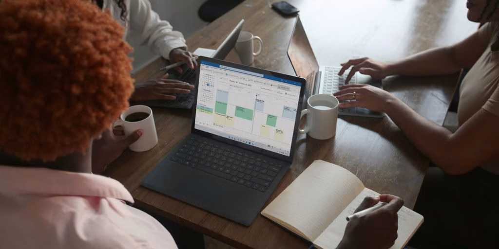 Three people in a meeting at a table discussing schedule on their Microsoft laptop
